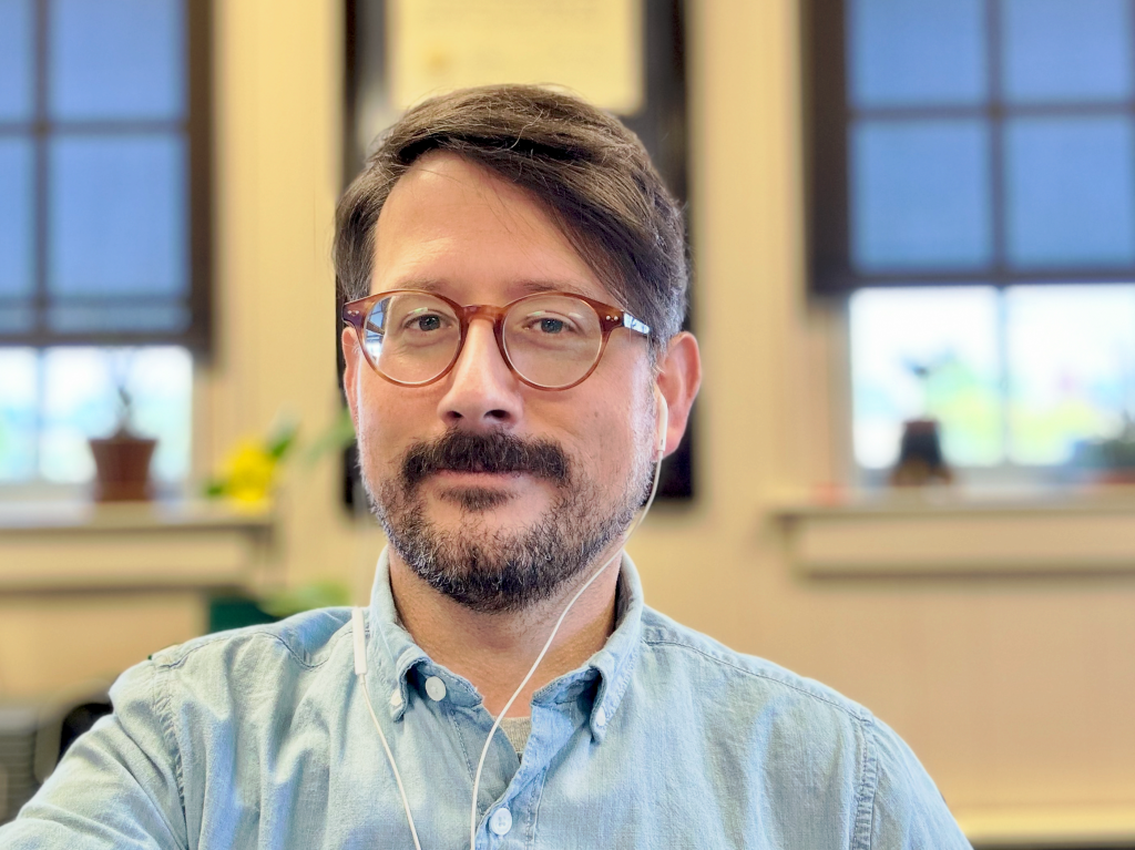 A close-up portrait of a man with glasses and a light blue shirt, sitting in an office environment. He has short, dark hair and is wearing earphones.